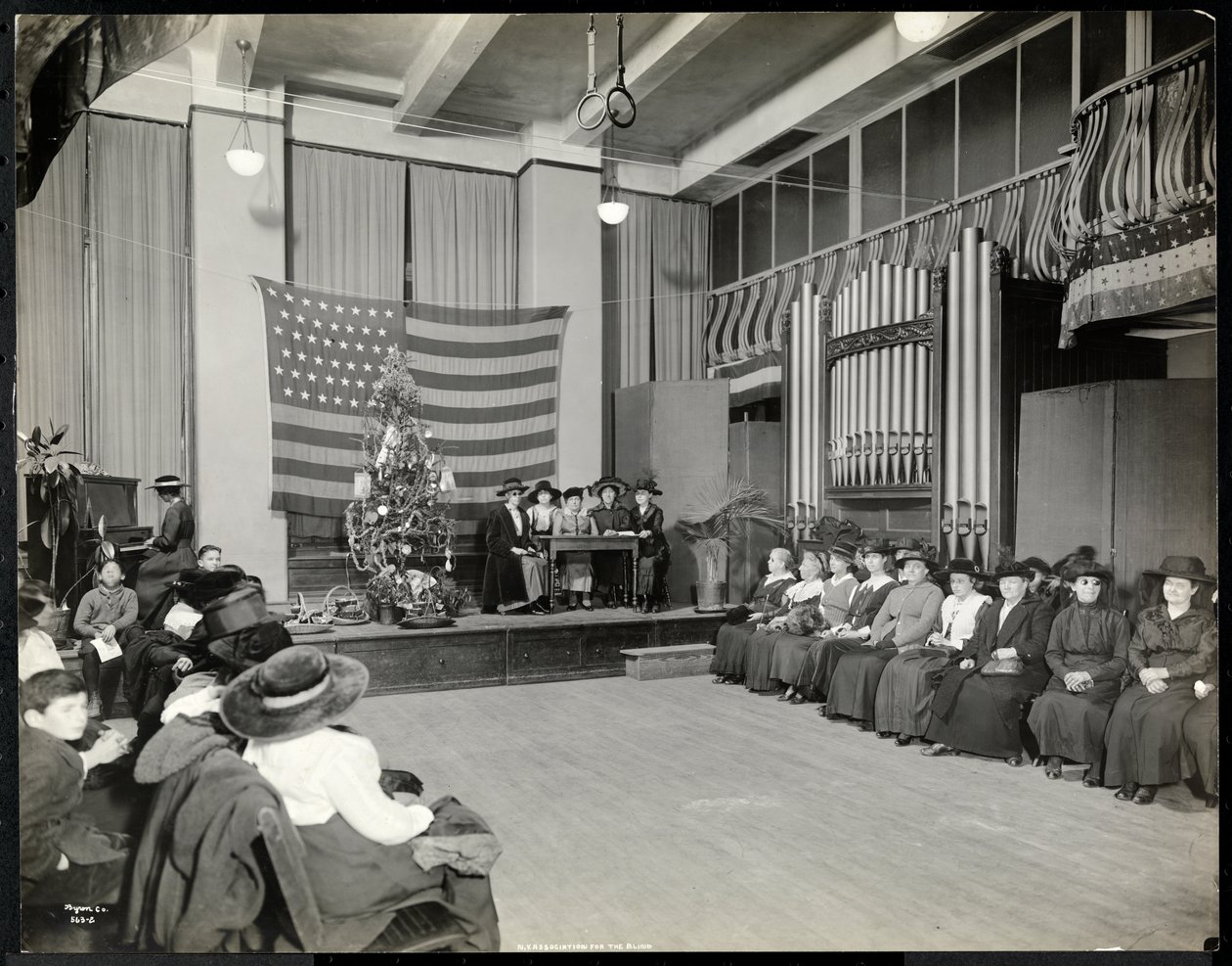 Personas en un auditorio en la Asociación para Ciegos de Nueva York con árbol de Navidad y personas posiblemente actuando en el escenario con órgano, 1920 de Byron Company