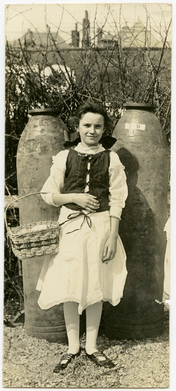 Fotografía sepia vertical de una niña con un vestido frente a dos grandes proyectiles de artillería. La niña sostiene una cesta en el codo derecho de Carl Michel