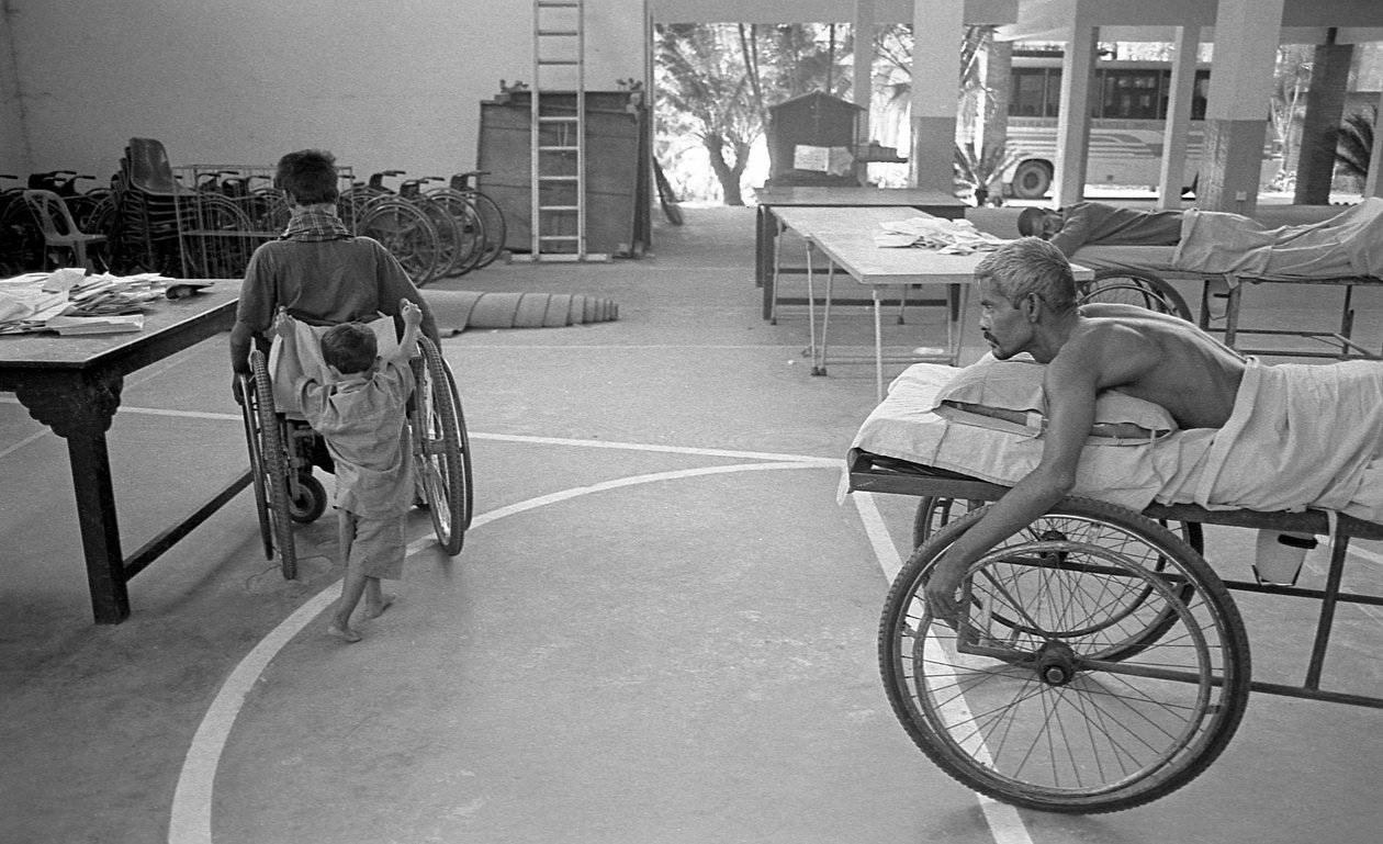 Un hombre con discapacidad física en una silla de ruedas es llevado por su hijo; otro hombre con la misma situación lo observa en el Centro para la Rehabilitación de los Paralíticos, CRP en Savar, Dhaka, Bangladesh. 2005. de Unbekannt Unbekannt